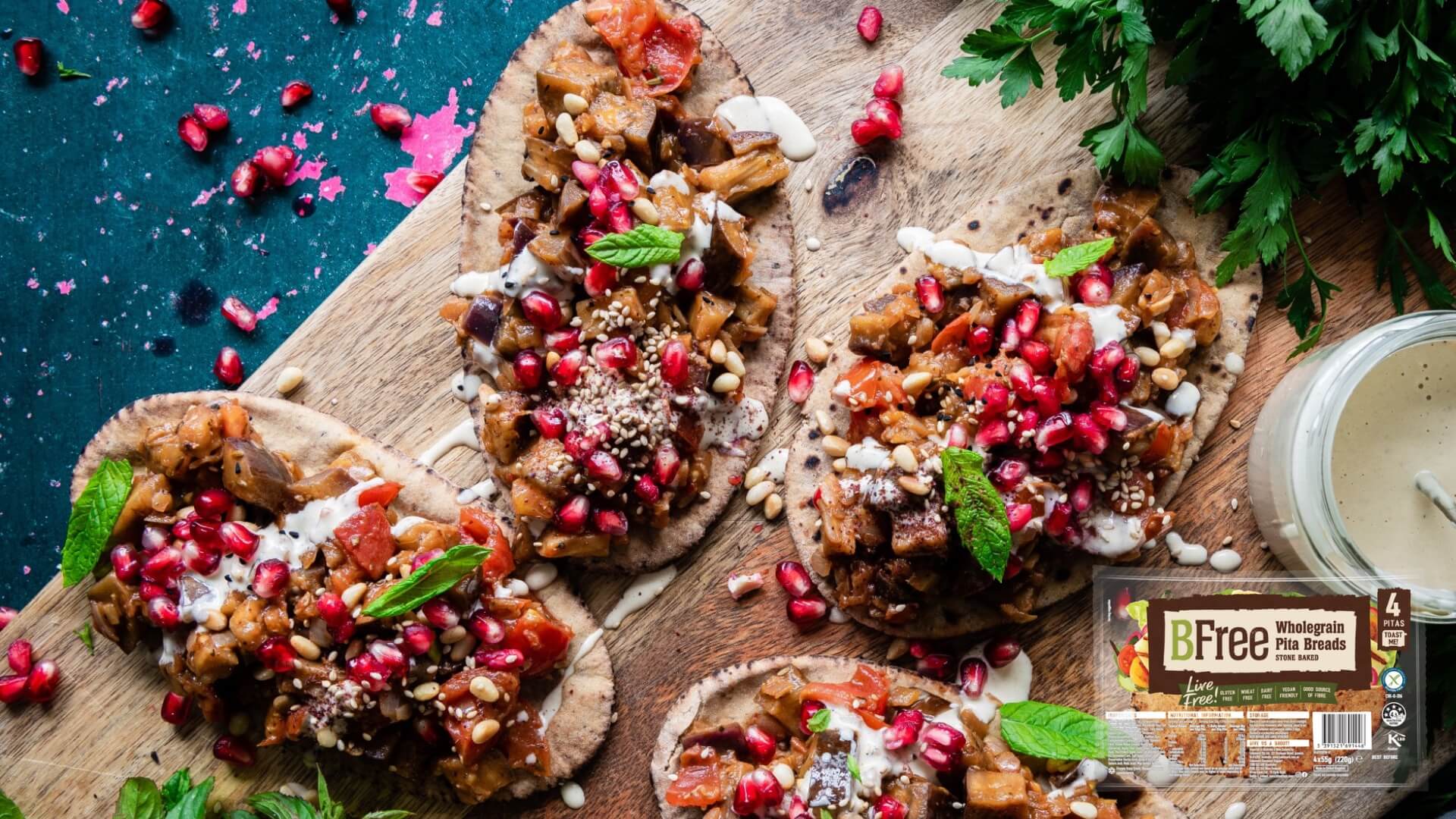 BFree gluten-free stone-baked pita bread. Image of pita breads on a serving board topped with spiced eggplant and pine nuts, pomegranate seeds and mint leaves.