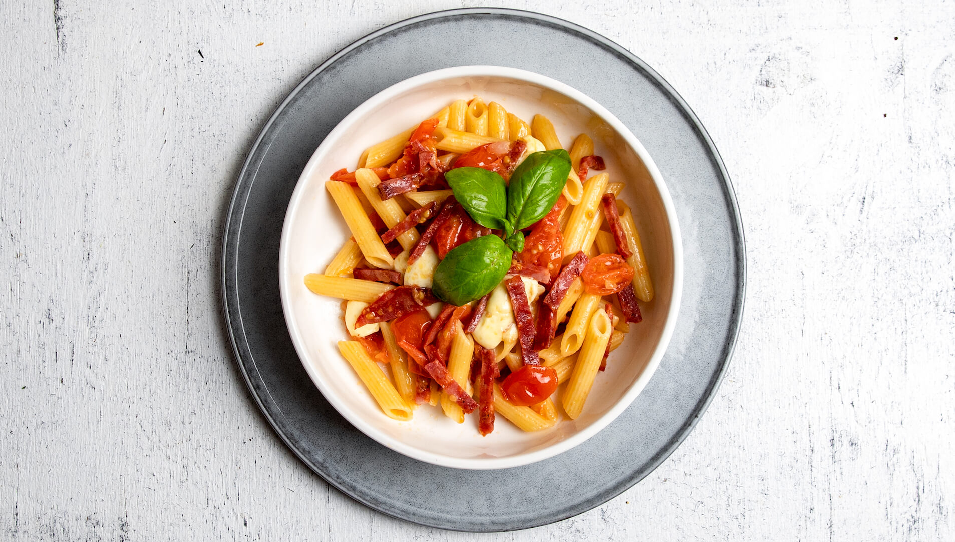 A bowl of penne piled with crispy salami, Roma tomatoes and bocconcini, topped with a sprig of fresh basil.