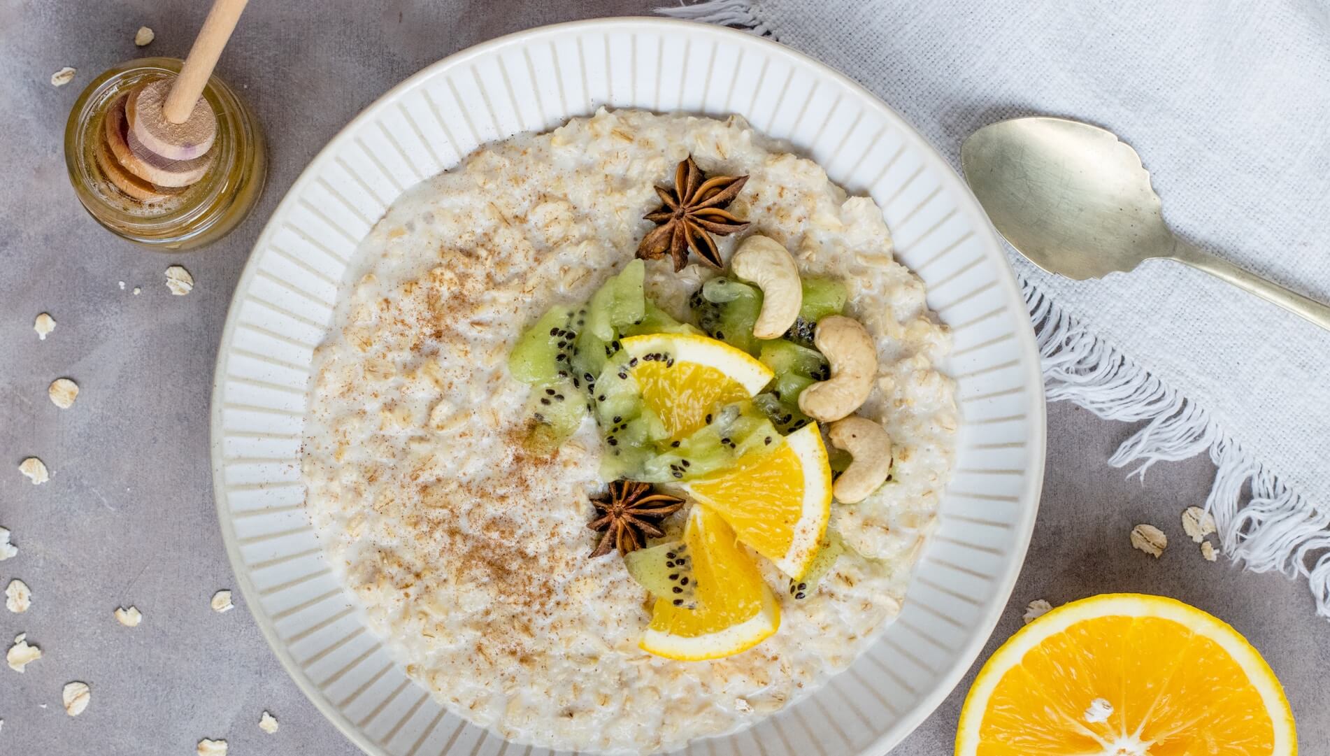 Are oats gluten free? A bowl of oats topped with citrus slices, kiwi, cashews and star anise with honey on the side.